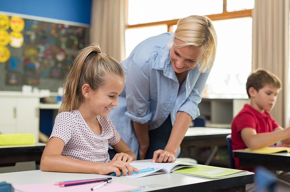 Happy teacher helping young girl with study in classroom. Teacher teaching to smiling schoolgirl at elementary school. Diligent child sitting at school desk with tutor.